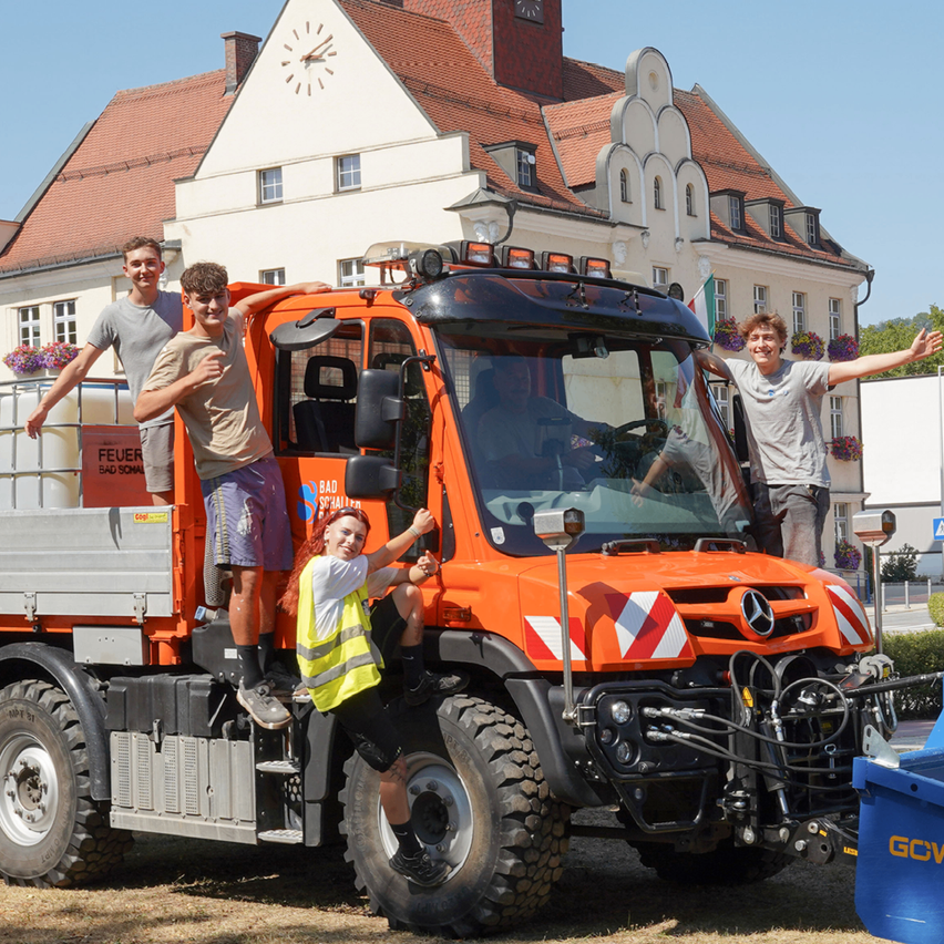 Four individuals stand on an orange Mercedes-Benz truck in front of a building with a clock tower.