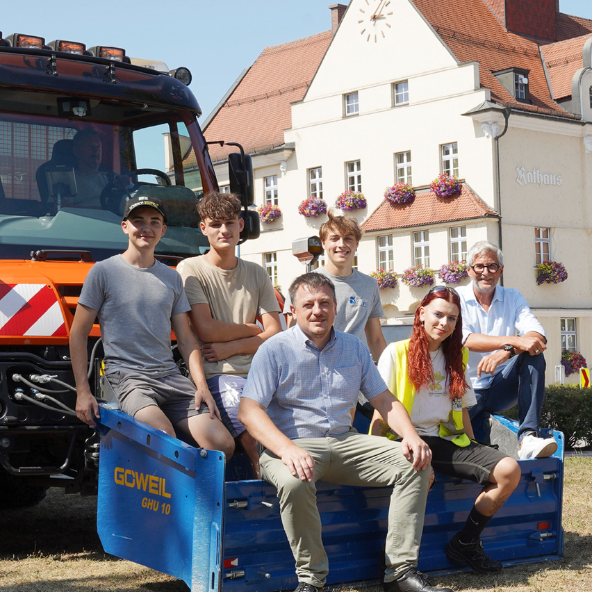 A group of people pose for a photo in front of a building with a clock tower and a truck with the text 'GCWEIL GHU 10'.