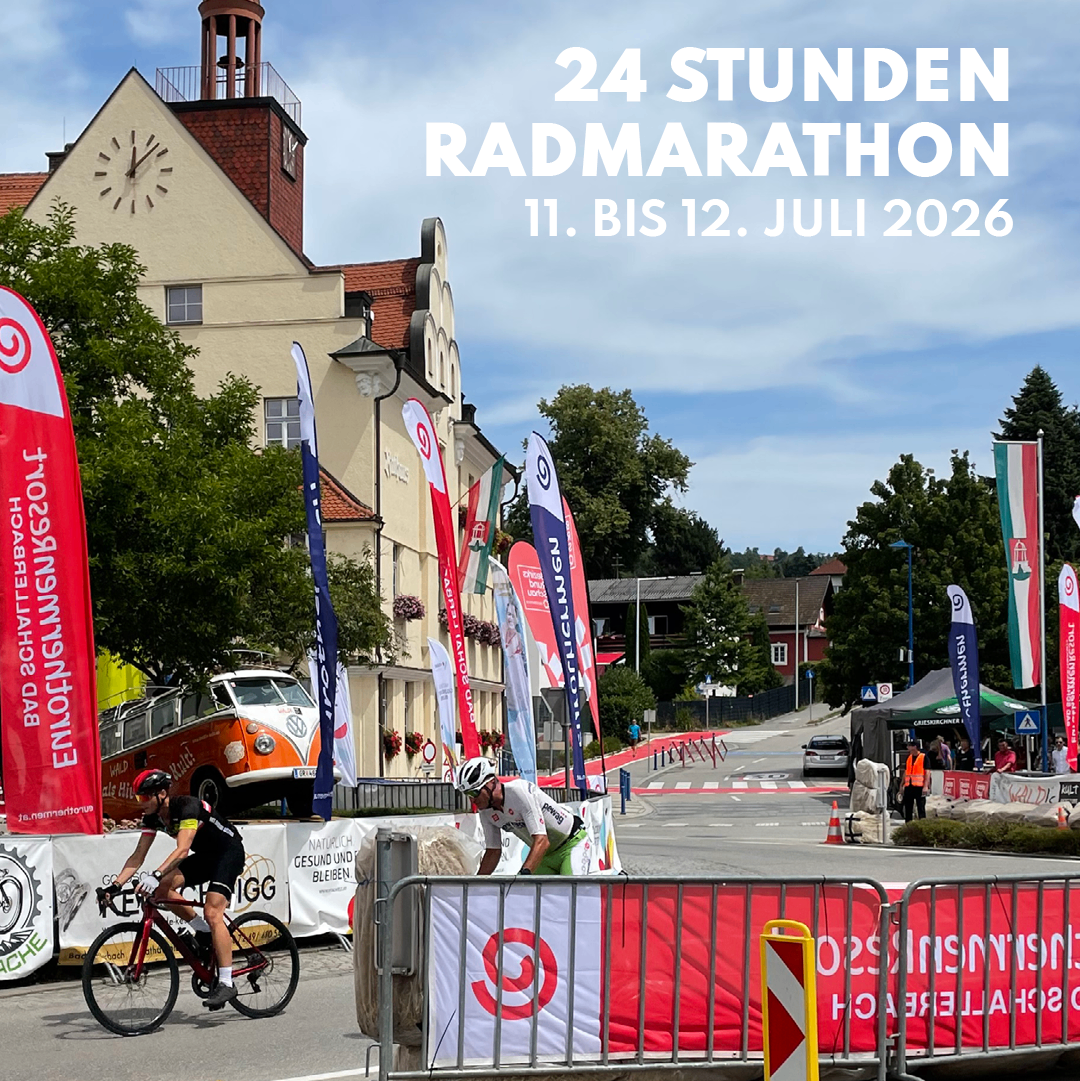 Zwei Radfahrer fahren im 24 Stunden Radmarathon, eine Veranstaltung vom 11. bis 12. Juli 2026. Sie fahren an Bannern und einem Gebäude mit Turm und Uhr vorbei.
