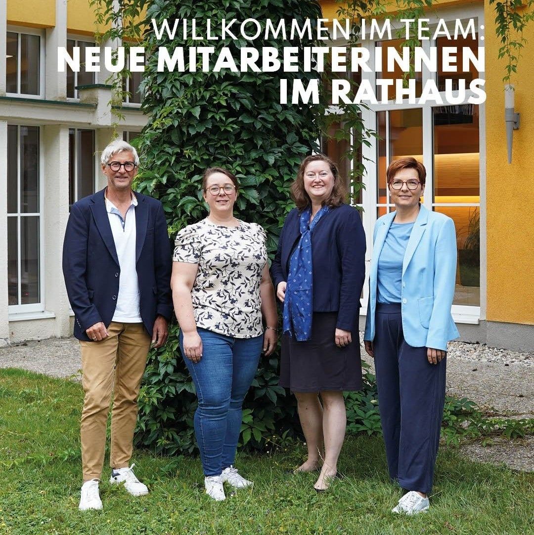 Fünf Personen stehen vor einem Gebäude mit gelber Wand und weißen Fenstern. Der Text lautet 'Willkommen im Team: Neue Mitarbeiterinnen im Rathaus'. Die Personen lächeln und scheinen für ein Foto zu posieren.