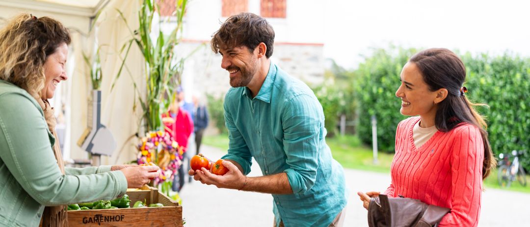 Ein Mann in einem blauen Hemd hält Tomaten in einem Gemüsemarkt. Neben ihm lächelt eine Frau in einem roten Pullover. Hinter ihnen greift eine Person nach grünem Gemüse.