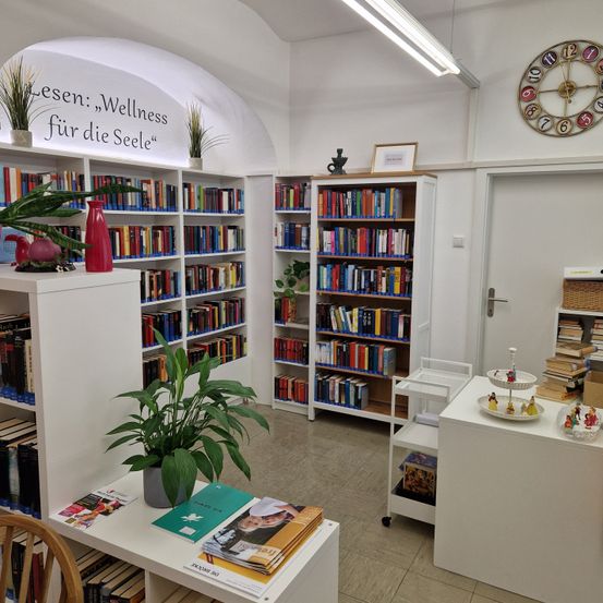 Eine Bibliothek voller Bücher auf weißen Regalen, eine große Pflanze in der Mitte und eine Uhr an der Wand. Ein Tisch mit Büchern und Figuren steht in der Nähe der Tür.