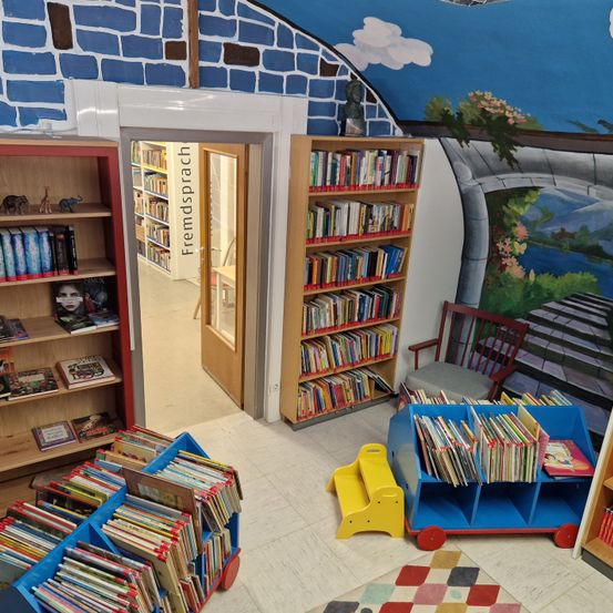 Eine gemütliche Kinderbibliothek mit einem Wandbild eines Hauses, ausgestattet mit Bücherregalen, einem blauen Bücherwagen und einem roten Stuhl. Der Eingang ist mit Fremdsprache beschriftet.