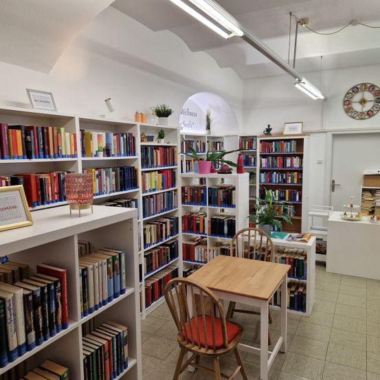 Eine kleine Bibliothek mit gewölbten Decken, gefüllt mit Büchern auf Regalen. Ein Holztisch mit Stühlen steht in der Mitte. Topfpflanzen und eine Uhr schmücken den Raum.