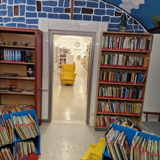 Eine Bibliotheksinnenansicht mit Regalen voller Bücher, einer Büste auf dem rechten Regal und einem gelben Stuhl in der Mitte, sowie einer blauen Ziegelsteinwand.