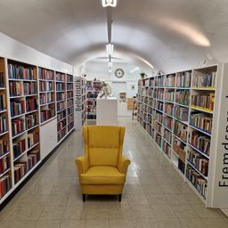 Ein gelber Sessel steht in der Mitte eines Bibliotheksflurs, mit Bücherregalen auf beiden Seiten, beschriftet mit 'Belletristik' und 'Fremdsprachen'.