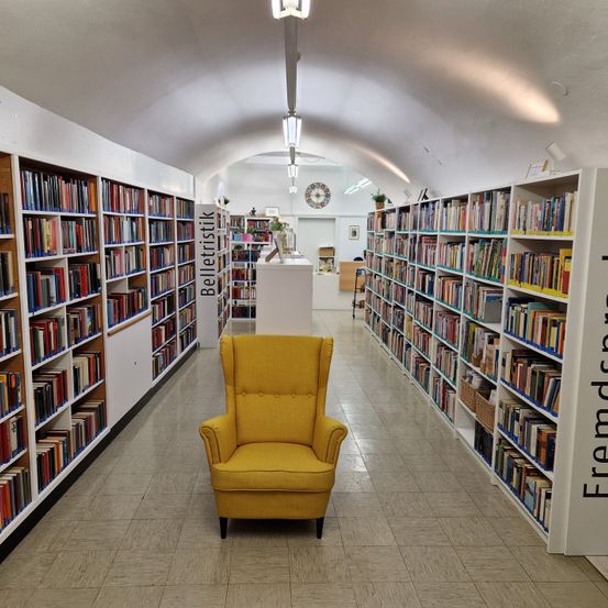 Ein gelber Sessel steht in der Mitte eines Bibliotheksflurs, mit Bücherregalen auf beiden Seiten, beschriftet mit 'Belletristik' und 'Fremdsprachen'.