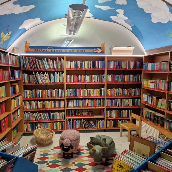 Eine Kinderbibliothek voller Bücher, mit einer Wolkendecke, Leseecken mit Plüschtieren und einer Vielzahl von Büchern auf Holzregalen.