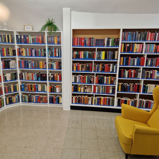 Innenansicht einer Bibliothek mit weißen Regalen voller Bücher, einem gelben Sessel in der Ecke und einer Pflanze auf dem obersten Regal.