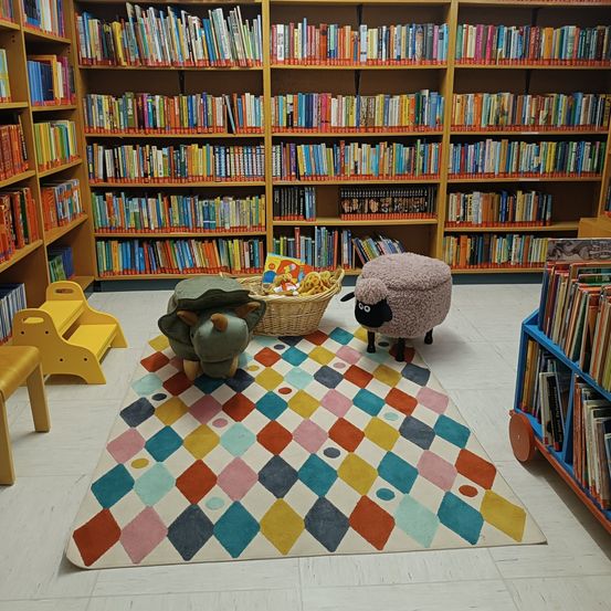 Ein farbenfroher Teppich liegt auf dem Boden einer Bibliothek. Gefüllte Tiere und ein Korb sitzen auf dem Teppich. Mit Büchern gefüllte Regale umgeben den Bereich. Ein gelber Stuhl und ein blaues Bücherregal sind sichtbar.