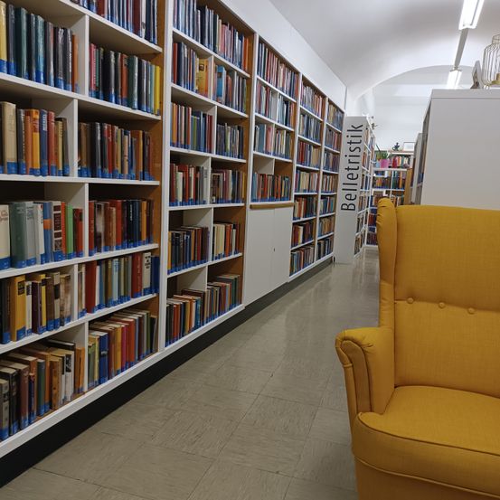 Ein Bibliotheksgang mit weißen Regalen, gefüllt mit Büchern. Ein gelber Sessel befindet sich auf der rechten Seite. Die Regale haben blaue Aufkleber. Der Gang ist gut beleuchtet.