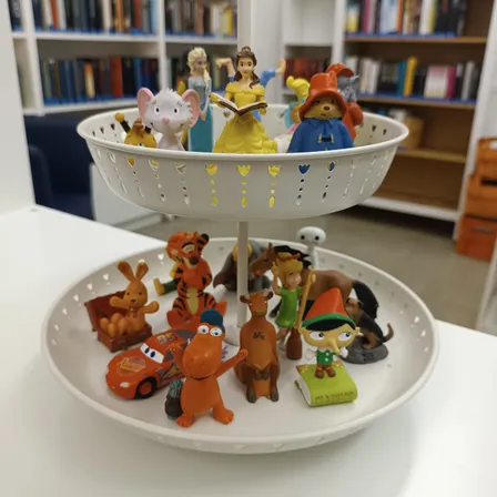 Ein zweistöckiger Ständer zeigt verschiedene Figuren auf einem weißen Tisch. Auf der oberen Ebene befinden sich Charaktere wie Belle, eine Fee und eine Prinzessin. Die untere Ebene hält eine Mischung aus Figuren, darunter ein Auto, ein Elefant und ein Hase.
