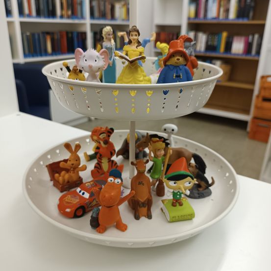 Ein zweistöckiger Ständer zeigt verschiedene Figuren auf einem weißen Tisch. Auf der oberen Ebene befinden sich Charaktere wie Belle, eine Fee und eine Prinzessin. Die untere Ebene hält eine Mischung aus Figuren, darunter ein Auto, ein Elefant und ein Hase.