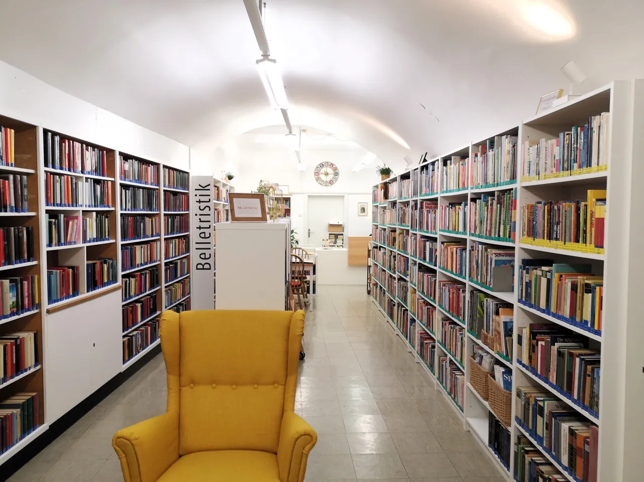 Eine Bibliothek mit Bücherregalen, einem gelben Stuhl und einer Uhr an der Wand.