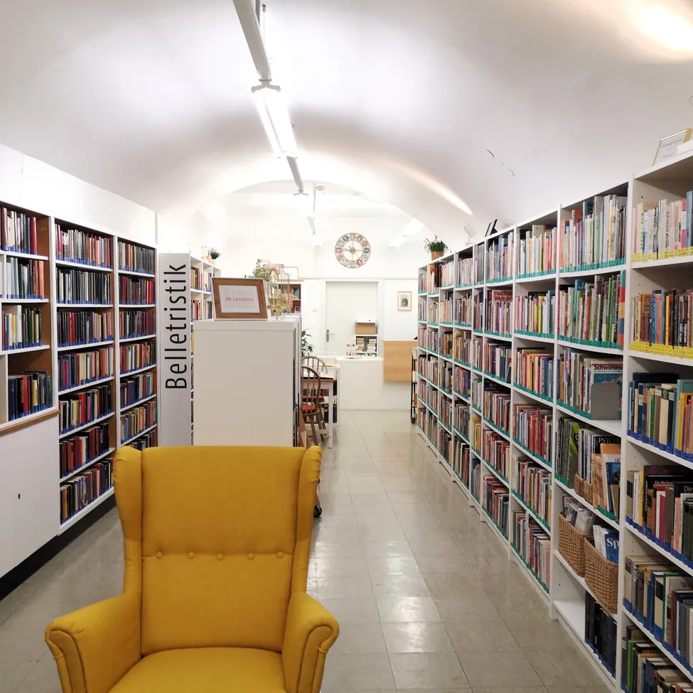Eine Bibliothek mit Bücherregalen, einem gelben Stuhl und einer Uhr an der Wand.