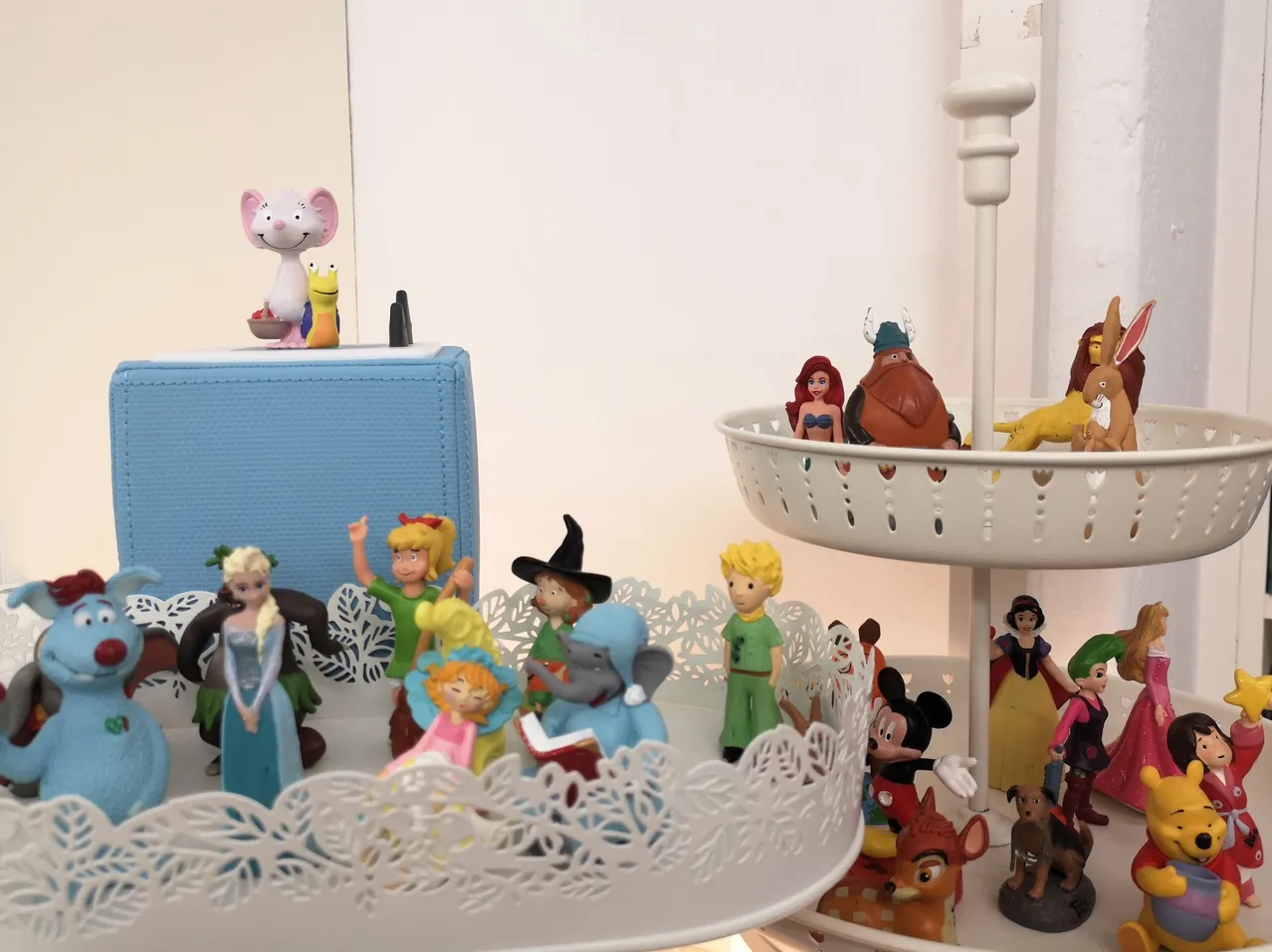 Eine Ausstellung verschiedener animierter Figuren, einschließlich Disney-Charaktere und anderer, auf einem zweistöckigen Ständer angeordnet. Oben sitzt eine Maus-Figur auf einer blauen Schachtel.