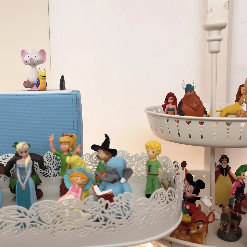 Eine Ausstellung verschiedener animierter Figuren, einschließlich Disney-Charaktere und anderer, auf einem zweistöckigen Ständer angeordnet. Oben sitzt eine Maus-Figur auf einer blauen Schachtel.