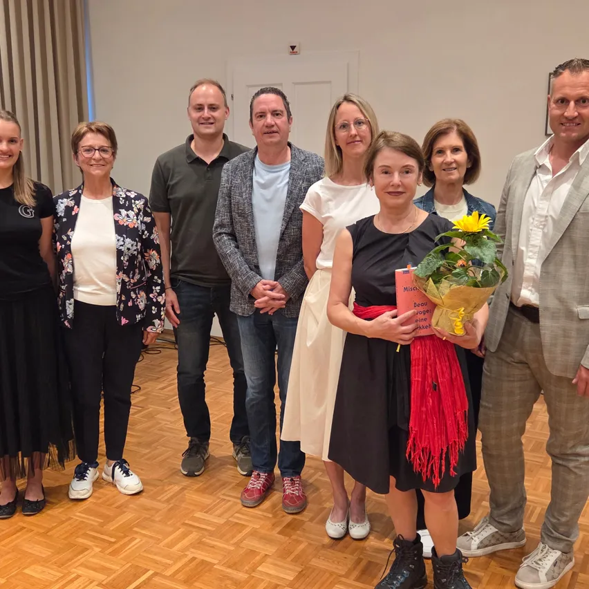 Eine Gruppe von Menschen steht in einem Raum mit Holzboden. Eine Frau hält einen Blumenstrauß und ein Buch in der Hand. Die anderen sehen sie an.