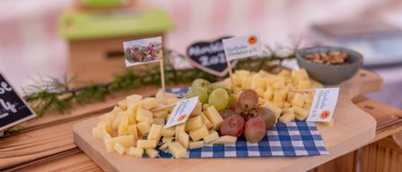 Ein Holztisch trägt ein Käsebrett mit verschiedenen Käsesorten und Trauben, verziert mit grünen und weißen Karten. Im Hintergrund befinden sich eine Holzbox und eine Tafel.