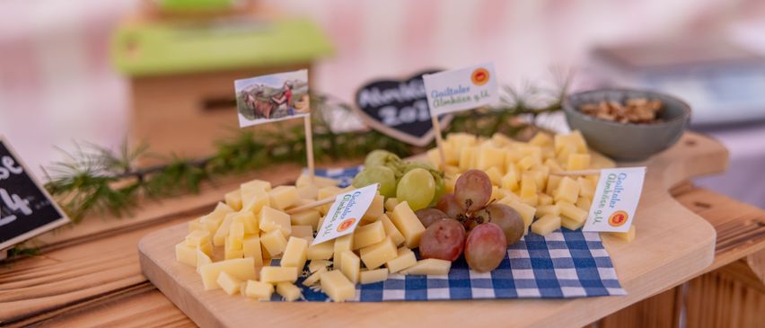 Ein Holztisch trägt ein Käsebrett mit verschiedenen Käsesorten und Trauben, verziert mit grünen und weißen Karten. Im Hintergrund befinden sich eine Holzbox und eine Tafel.