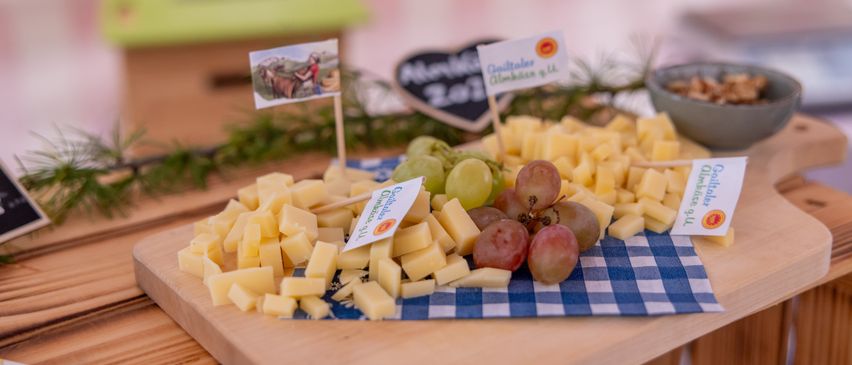 Ein Holzschneidebrett mit Käsewürfeln und Trauben auf einem blau-weiß karierten Tuch, mit einem Schild, das 'Almkäserei' sagt. Dahinter ist eine Tafel mit der Nummer '201'.