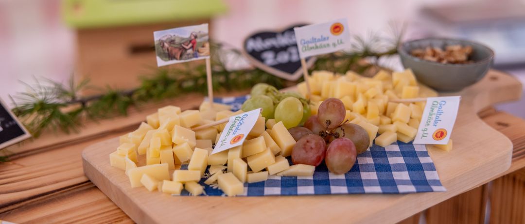 Ein Holzschneidebrett mit Käsewürfeln und Trauben auf einem blau-weiß karierten Tuch, mit einem Schild, das 'Almkäserei' sagt. Dahinter ist eine Tafel mit der Nummer '201'.