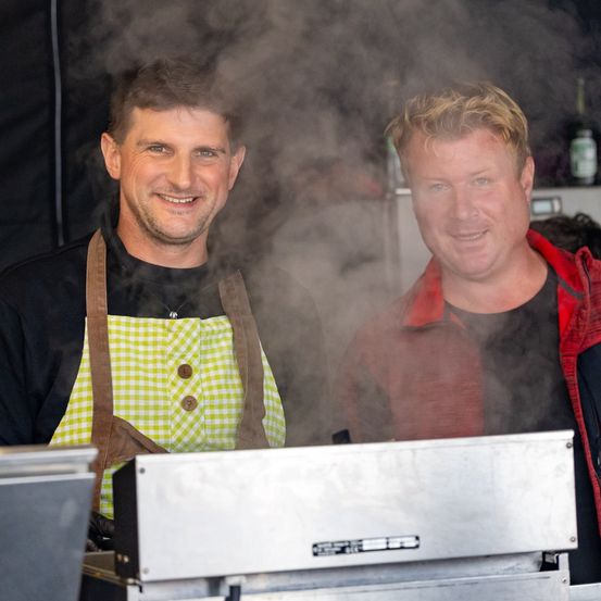 Zwei Männer stehen lächelnd vor einem dampfenden Imbisswagen. Einer trägt eine grün-gelb karierte Schürze, der andere eine rot-schwarze Jacke. Rauch steigt vom Wagen auf.
