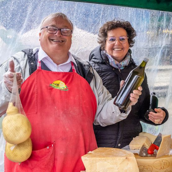 Ein Mann in einer roten Schürze hält gelben Käse und eine Flasche Wein und steht neben einer lächelnden Frau, die eine Flasche hält. Sie sind von verschiedenen Käsesorten umgeben.