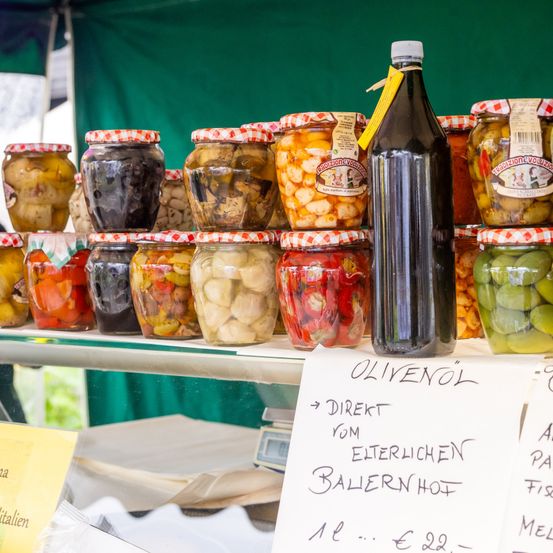 Ein Tisch zeigt mehrere Gläser mit eingelegtem Gemüse und Oliven unter einem grünen Zelt. Eine Flasche Olivenöl steht prominent in der Mitte. Ein Schild liest 'Olivenol Direkt vom Eiterlichen Ballernhof'.