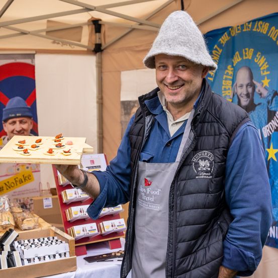 Ein Mann lächelt und hält eine Platte mit kleinen Gerichten mit roten Toppings. Er trägt einen Hut und eine Schürze mit dem Logo 'Slow Food Travel'. Hinter ihm steht ein Zelt mit einem Schild und einem Banner mit dem Gesicht eines Mannes.