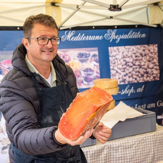 Ein Mann in einer schwarzen Jacke und Schürze lächelt und hält ein großes Stück Käse unter einem Zelt mit einem Banner, das 'Mediterrane Spezialitäten' steht.