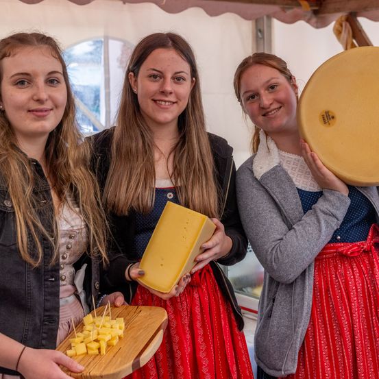 Drei Frauen stehen lächelnd nebeneinander und halten Käse. Die Frau auf der linken Seite hält ein Holzbrett mit Käsewürfeln. Die Frau in der Mitte hält ein großes Stück Käse. Die Frau auf der rechten Seite hält einen runden Käse mit einem Etikett. Sie befinden sich in einem Zelt.