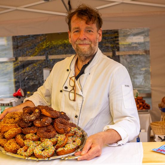 Ein Koch in weißer Uniform hält eine Platte mit Donuts und lächelt in die Kamera. Hinter ihm ein Zelt mit einer abstrakten Malerei und einem Korb mit Brot.