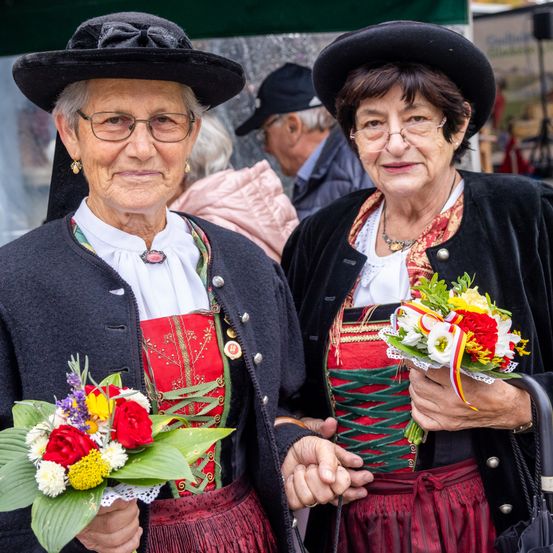 Zwei ältere Frauen in traditionellen Kostümen halten Blumensträuße. Sie stehen zusammen und lächeln, mit einem verschwommenen Hintergrund von Menschen.