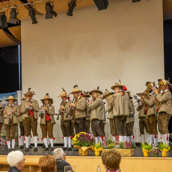 Eine Gruppe von Musikern in traditionellen Uniformen spielt auf der Bühne mit verschiedenen Instrumenten. Die Bühne ist mit Blumen geschmückt, und eine weiße Leinwand steht dahinter. Das Publikum sitzt vor der Bühne.