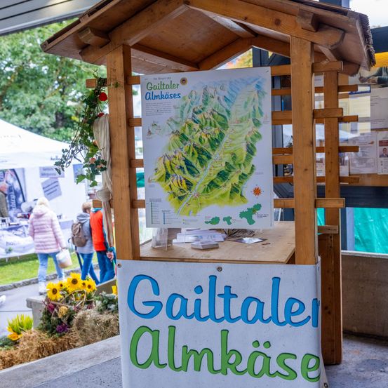 Ein hölzerner Stand zeigt eine Karte der Gailtaler Almkaeses mit einem grünen Dach und Blumen auf dem Boden. Im Hintergrund gehen Menschen.