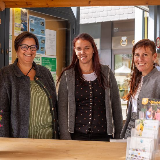 Drei lächelnde Frauen stehen hinter einem Holzschreibtisch mit Blumen. Die Frau auf der linken Seite trägt eine Brille und ein grünes Kleid. Die mittlere Frau hat einen grauen Cardigan und eine Halskette. Die rechte Frau trägt ein weißes Hemd und hat lange Haare. Hinter ihnen ist eine Glastür mit Plakaten.