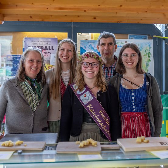 Fünf lächelnde Personen, darunter zwei Frauen in traditioneller Kleidung, stehen vor einer Glasvitrine mit Käse. Dahinter sind Poster an der Wand angebracht.