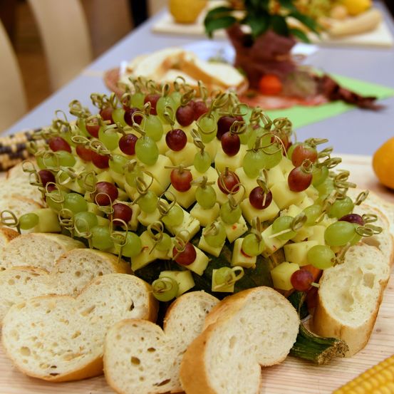 Auf einem Tisch steht ein Käse- und Trauben-Vorspeisenteller mit Zahnstochern und einem Maiskolben an der Seite. Im Hintergrund befinden sich Brot und eine weitere Speiseplatte.