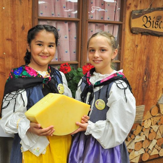 Zwei junge Mädchen in traditionellen Kostümen stehen vor einem Holzgebäude und halten ein großes Stück Käse. Hinter ihnen befindet sich ein Fenster mit einem rot-weiß karierten Vorhang und ein Schild mit dem Wort 'Bisca'.
