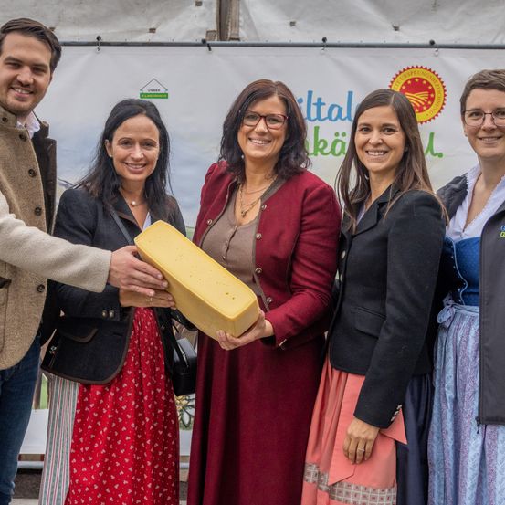 Eine Gruppe von Menschen steht vor einem weißen Banner mit Logo. Sie lächeln und posieren für ein Foto. Die Frau in der Mitte hält einen gelben Käse. Die Frau auf der linken Seite trägt ein rotes Kleid. Die Frau auf der rechten Seite trägt eine Brille.