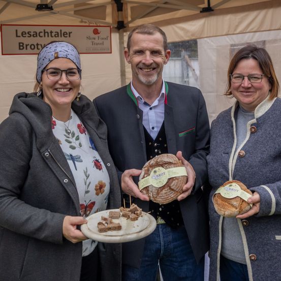 Drei Personen stehen unter einem Zelt und halten Scheiben Brot in den Händen. Eine Frau auf der linken Seite hält einen Teller mit Brotstücken, während der Mann in der Mitte ein ganzes Brot hält. Die Frau auf der rechten Seite hält ein Brot mit einem Etikett. Hinter ihnen steht ein Schild mit der Aufschrift 'Lesachtaler Broty Slow Food Travel'.