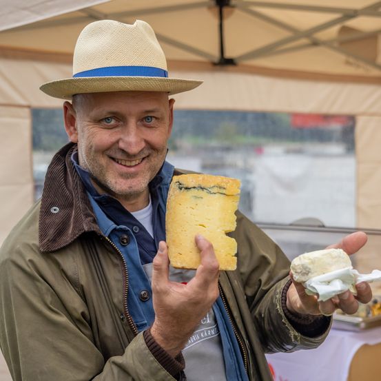 Ein Mann in Hut und Jacke hält ein Stück Käse und eine kleine Menge Käse in den Händen und lächelt zur Kamera.