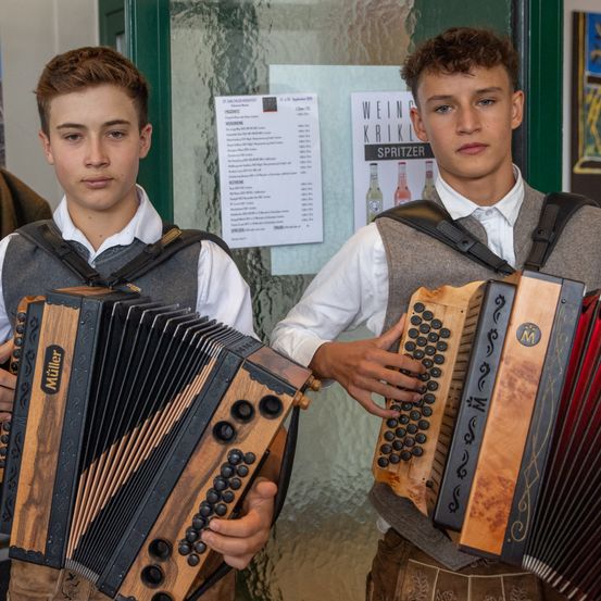 Zwei junge Männer in traditioneller deutscher Kleidung stehen mit Akkordeons in der Hand. Einer trägt eine Weste und der andere einen Rucksack. Sie stehen vor einer Glastür mit einer Speisekarte.