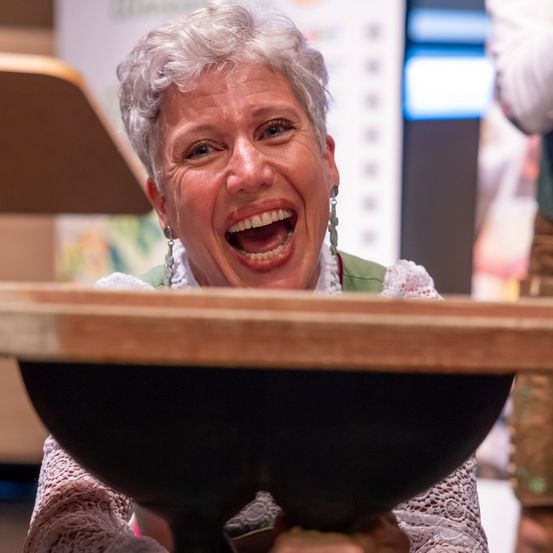 Eine Frau in einem rosa Kleid mit Spitzenärmeln hält einen großen schwarzen Topf über ihrem Kopf und lacht mit offenem Mund.