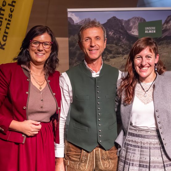 Vier Personen stehen lächelnd vor einer Kulisse mit einer Berglandschaft. Zwei Frauen und ein Mann tragen traditionelle deutsche Kleidung.