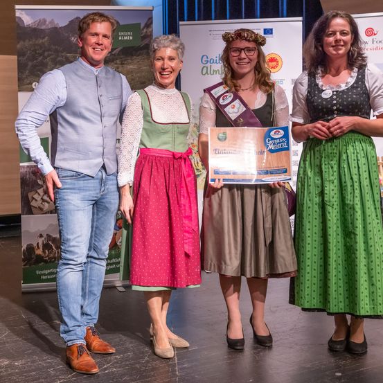 Vier Personen stehen lächelnd auf einer Bühne. Ein Mann in einem hellblauen Hemd und Jeans hält einen Preis in der Hand, neben ihm stehen zwei Frauen in traditioneller deutscher Kleidung und eine Frau mit Brille. Hinter ihnen sind Poster an der Wand angebracht.