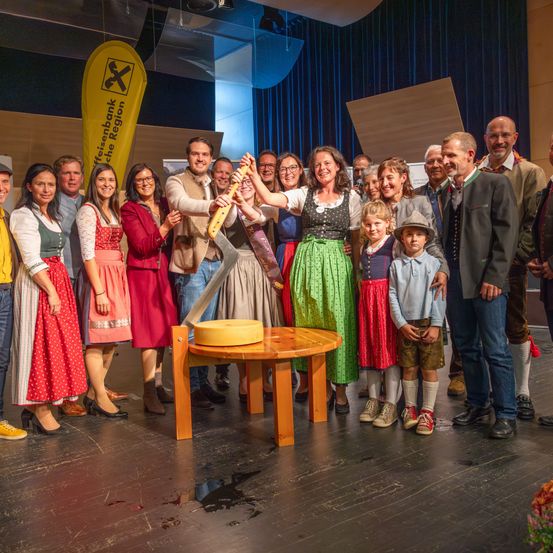 Eine Gruppe von Menschen in traditioneller bayerischer Kleidung steht um einen Holztisch mit einem großen Käserad und hält ein großes Messer, um es zu schneiden. Die Szene spielt auf einer Bühne mit einem gelben Banner und blauen Vorhängen im Hintergrund.