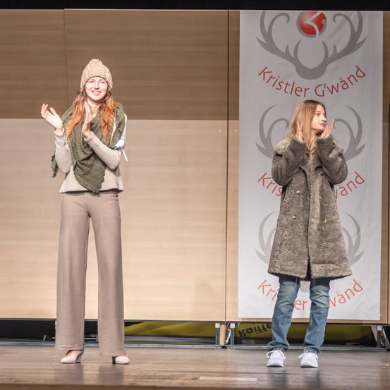 Zwei Frauen stehen auf der Bühne vor einem Banner mit der Aufschrift 'Kristler Gwand'. Die Frau auf der linken Seite trägt eine Mütze, einen Schal und hohe Schuhe. Die Frau auf der rechten Seite trägt einen Mantel und Jeans.