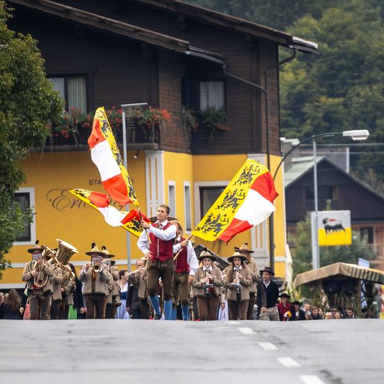 Ein Musikzug führt eine Parade mit zwei Fahnen an einem sonnigen Tag vor einem gelben Gebäude mit Balkonen und Blumen an.